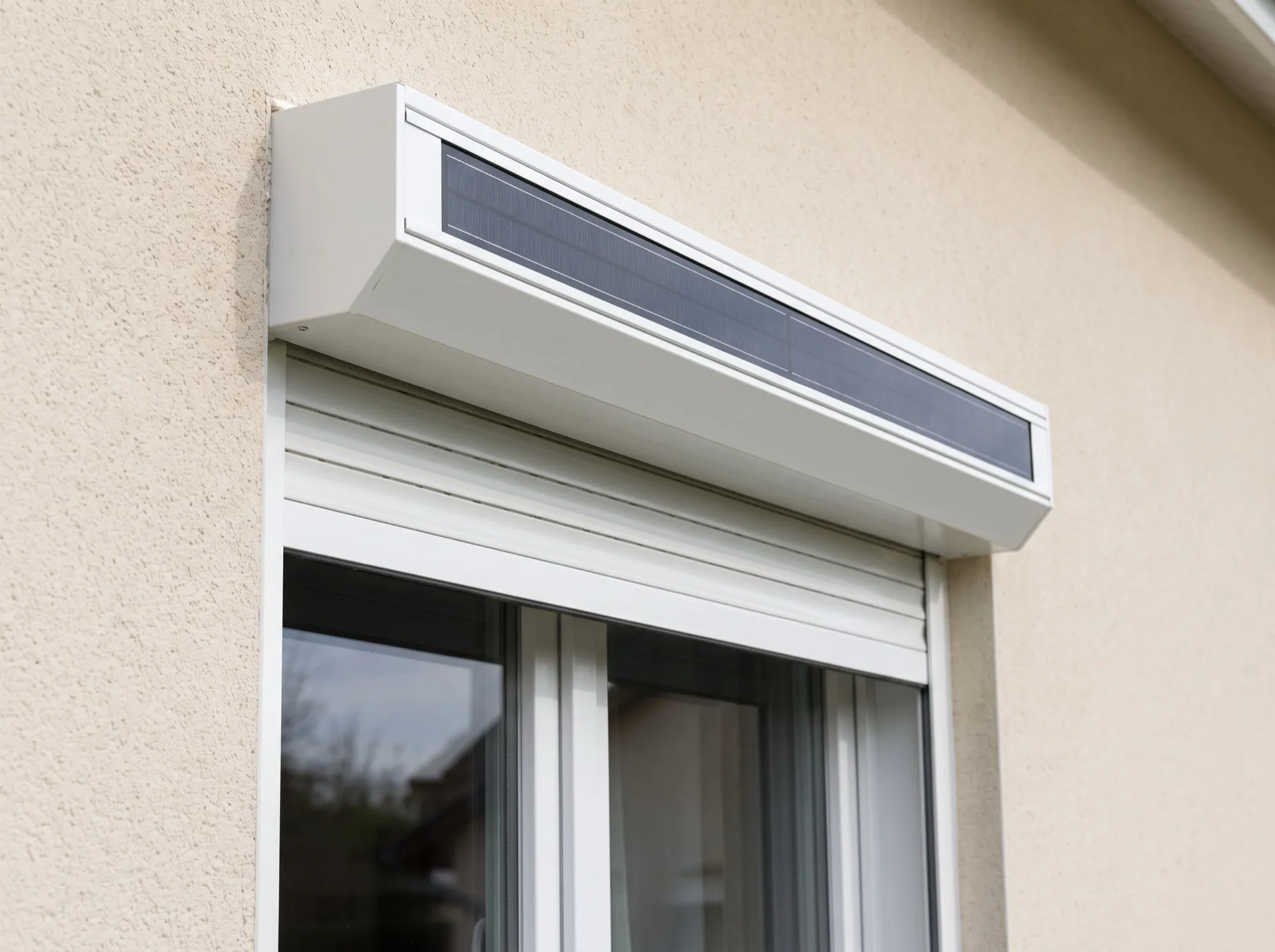 Gros plan sur un coffre de volet roulant blanc avec sa bande photovoltaïque intégrée, au-dessus d'une fenêtre PVC